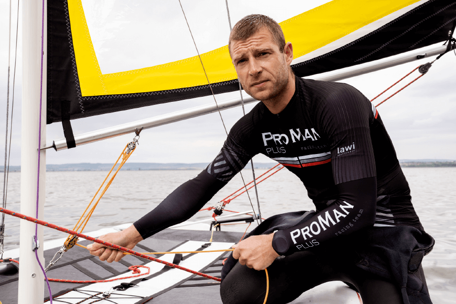Natural Pharmaceuticals ProMan® Plus Egy férfi sportöltönyben, „ProMan® Plus” és „racing team” logókkal egy hajóban vitorlázik, kezében a kormánykötelekkel. A háttérben egy nyugodt vízfelület és a horizont a szárazföld körvonalaival. A férfi koncentráltnak tűnik, és öltözéke egy profi regattán való részvételre utal.
